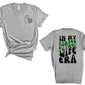 May include: Gray t-shirt with a green border patrol badge on the left chest and the text "In My Border Patrol Wife Era" in black and green on the back.