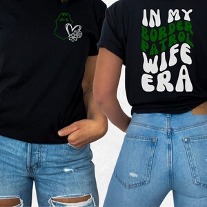 May include: Black t-shirt with a green outline of a badge and a heart with flowers on the left side. The back of the shirt has the text "IN MY BORDER PATROL WIFE ERA" in white with green outlines.