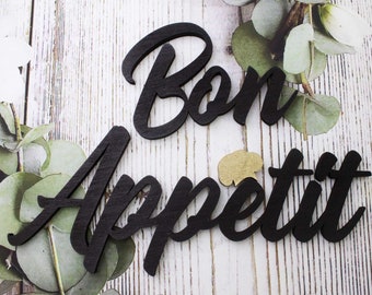 May include: Black wooden letters spelling "Bon Appetit" on a white wood background with green eucalyptus leaves.