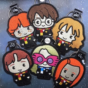 May include: Six embroidered keychains featuring characters from the Harry Potter series. The characters are Harry Potter, Ron Weasley, Hermione Granger, Luna Lovegood, Ginny Weasley, and Neville Longbottom. Each keychain is made of black leather with a gold chain and a small charm.
