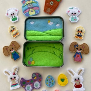 May include: A set of felt Easter-themed appliques in a tin box. The appliques include a house, a bunny, a chick, a lamb, a carrot, a bird, and eggs. The tin box has a green felt interior with a blue felt sky and clouds.