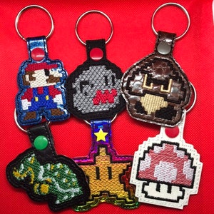 May include: A collection of six embroidered keychains featuring pixelated video game characters. The keychains include Mario, a ghost, a mushroom, a star, a turtle, and a red and white mushroom. Each keychain has a metal ring and a leather backing.