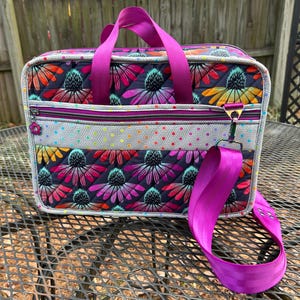 May include: A quilted tote bag with a floral print in shades of pink, purple, and green. The bag has a zippered pocket on the front and a detachable shoulder strap. The strap is a bright purple color.