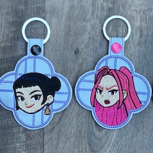 May include: Four embroidered keychains with a light blue quatrefoil design. Each keychain features a different cartoon character's face, with pink, black, and light brown hair. The keychains have a silver key ring attached.