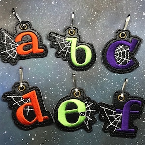 May include: Six Halloween-themed zipper pulls with letters A through F. Each pull features a black background with a white spiderweb design and a colorful letter in the center. The letters are orange, green, purple, orange, green, and purple.