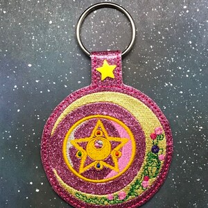 May include: Pink and gold glitter keychain with a crescent moon and a five-pointed star with a smaller moon inside. The star has five gems on the points.