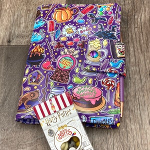 May include: A purple Harry Potter-themed journal with a colorful pattern of magical icons and treats, including a birthday cake and a box of Bertie Bott's Every Flavor Beans. The journal is closed with a snap closure.