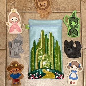 May include: A blue zippered pouch with a green and yellow embroidered image of the yellow brick road leading to the Emerald City.  The pouch is surrounded by eight embroidered characters from the Wizard of Oz, including Dorothy, the Scarecrow, the Tin Man, the Cowardly Lion, the Wicked Witch of the West, Glinda the Good Witch, and Toto the dog.