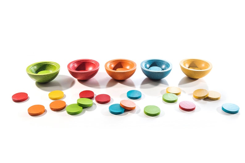 Peut inclure: Un ensemble de cinq bols en bois de diff&eacute;rentes couleurs, notamment vert, rouge, orange, bleu et jaune. Les bols sont dispos&eacute;s en rang&eacute;e et plusieurs petits disques en bois sont &eacute;parpill&eacute;s autour d'eux dans des couleurs assorties.