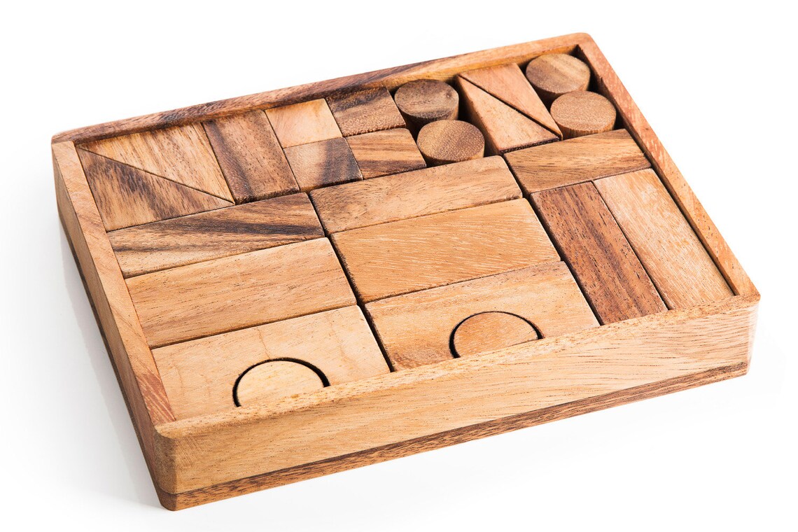 May include: A wooden box filled with various wooden blocks of different shapes and sizes. The blocks are arranged in a grid pattern and include squares, rectangles, triangles, and circles. The box is made of light brown wood and has a smooth finish.