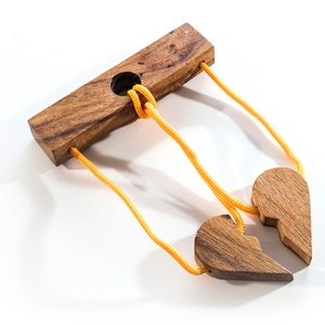 May include: A wooden puzzle with two heart-shaped pieces connected by yellow string. The string is threaded through a hole in a wooden block.