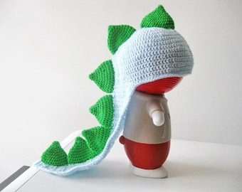 May include: A light blue crocheted hat featuring a row of bright green triangular spikes along the top and back, extending into a long tail-like section. The hat is displayed on a red and white figurine with a smooth, rounded surface.
