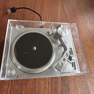 May include: Silver Crosley RSD3 portable turntable with a black record player and clear plastic cover. The turntable has a USB cable attached and sits on a wooden surface.