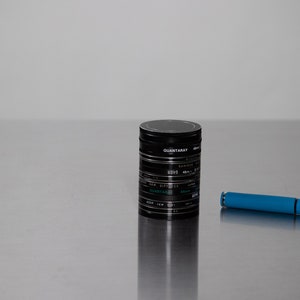 May include: A black cylindrical container with the text "QUANTA RAY" and "EDGE" printed on it. A blue pen is lying on a silver surface.