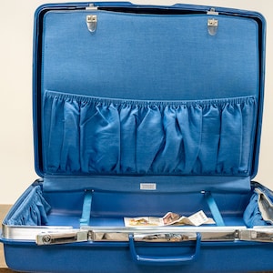 May include: A blue hard-sided suitcase with a silver handle and a blue fabric lining. The suitcase is open and has a zippered pocket on the inside.
