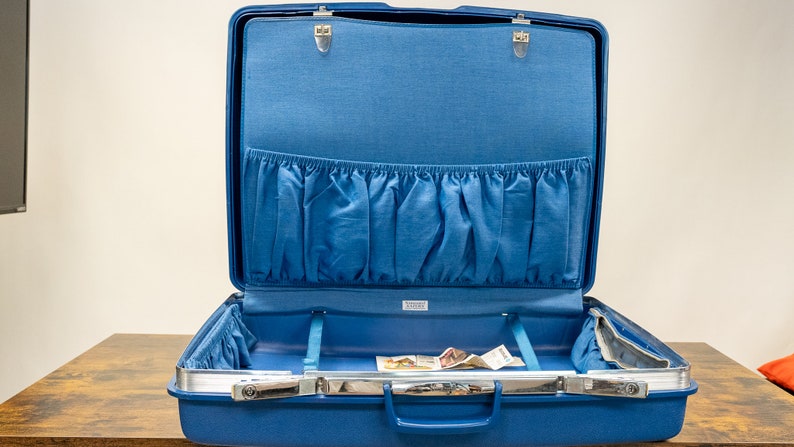 May include: A blue hard-sided suitcase with a silver handle and a blue fabric lining. The suitcase is open and has a zippered pocket on the inside.