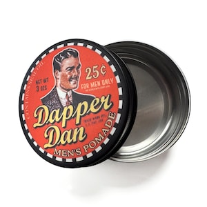 May include: Open tin of Dapper Dan Men's Pomade. The lid is red with a vintage illustration of a man in a suit and tie. Text on the lid reads "Dapper Dan Men's Pomade" and "25¢ for men only". The tin contains 3 oz of product.
