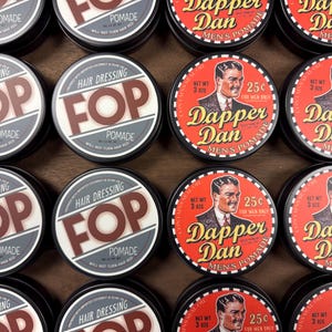 May include: A collection of vintage-style hair pomade tins. Some tins feature the text "FOP Pomade" in a retro font, while others display "Dapper Dan Men's Pomade" with a portrait illustration. The tins are arranged in a wooden box.