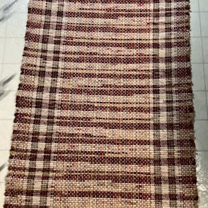 May include: A rectangular woven rug with a plaid pattern in shades of burgundy, cream, and brown. The rug has a fringed edge at one end. The weave is tight, creating a durable texture. This rug is suitable for a hallway or entryway.
