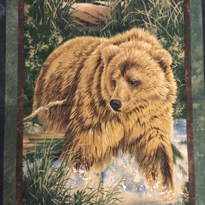 May include: A fabric panel featuring a detailed illustration of a brown grizzly bear walking through water. The bear has thick fur and is surrounded by green grass and a forest background. The panel is framed with a dark green border.