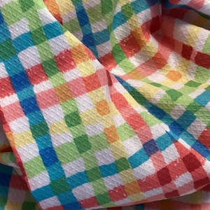 May include: Close-up of a colorful fabric with a gingham pattern. The fabric features a repeating checkered design in various colors, including red, blue, green, yellow, and orange, set against a white background. The fabric is textured and appears to be soft.