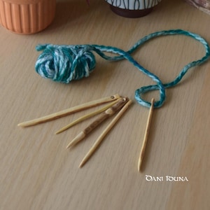 May include: A ball of green and white yarn with a long green and white cord, and five wooden knitting needles on a light wood surface.
