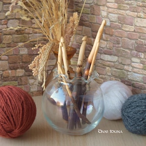 Op de afbeelding: Een heldere glazen kom met een gegolfde rand bevat verschillende houten haaknaalden. Links ligt een grote bol roestkleurig garen, rechts witte en grijze garenbollen. Gedroogde tarwe staat op de achtergrond.