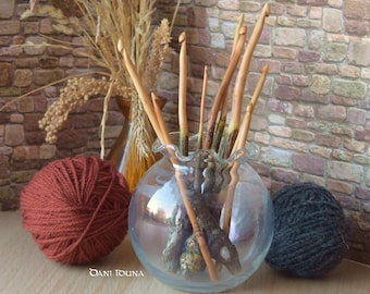 May include: A collection of light brown wooden crochet hooks with textured, bark-like handles stored in a clear, ruffled-rim glass bowl, accompanied by a ball of rust-colored yarn and a ball of dark gray yarn, with the text Dani Iduna visible.