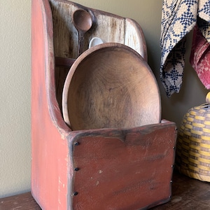 May include: A red wooden storage box with a distressed finish and black metal accents. The box has a shelf and a bowl inside. The box is sitting on a wooden table.