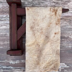 May include: A rustic wooden towel rack with a distressed red paint finish. A white towel with brown stains hangs on the rack.