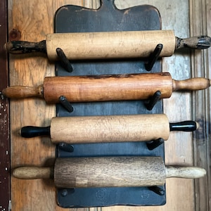 May include: Four wooden rolling pins with black handles are hanging on a black painted wooden rack. The rack is mounted on a wooden wall.