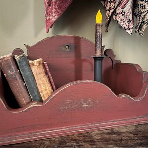 May include: A distressed red wooden shelf with a candle and several antique books. The shelf has a scalloped design and is painted in a rustic style. The candle is black with a yellow flame.
