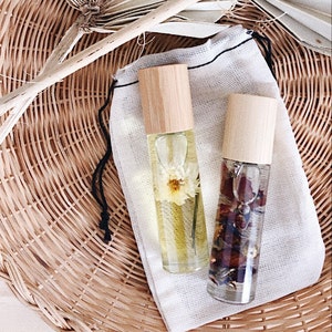 Puede incluir: Dos botellas roll-on de vidrio transparente con tapas de madera, llenas de líquido y elementos botánicos. Una contiene una flor blanca, la otra pétalos secos. Descansan sobre una bolsa de tela blanca, sobre una cesta de mimbre tejida.