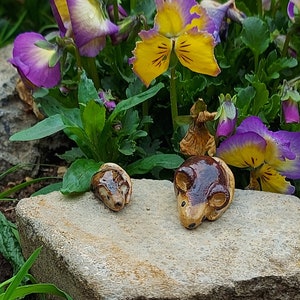 May include: Two small ceramic brown and white mice figurines on a grey rock with purple and yellow flowers in the background.