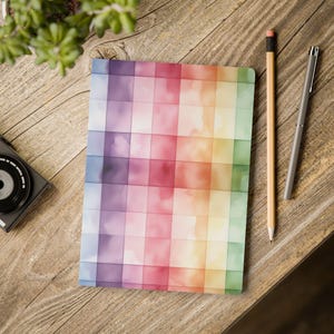 May include: A notebook with a rainbow grid design in purple, red, yellow, and green hues. A pencil and a pen lie beside the notebook on a wooden surface. A small camera and a plant are also visible.