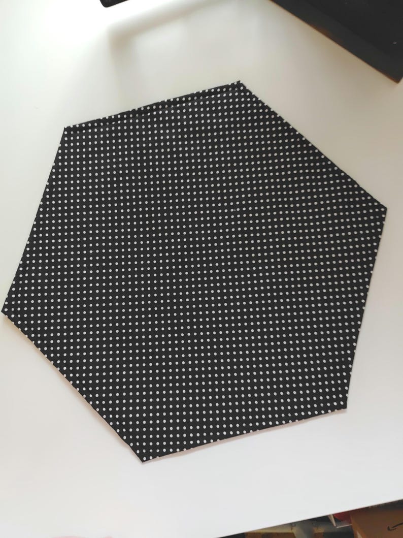 K&ouml;nnte beinhalten: Ein schwarzer, sechseckiger Stoffuntersetzer mit wei&szlig;em Tupfenmuster. Der Untersetzer besteht aus einem weichen Material und sch&uuml;tzt Oberfl&auml;chen vor Hitze und Feuchtigkeit. Das Design ist schlicht und modern.