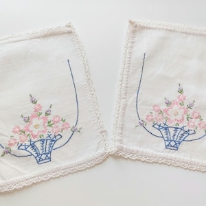 May include: Two white embroidered linen napkins with a floral basket design. The napkins feature pink and purple flowers in a blue basket, outlined with blue stitching. The edges are trimmed with a delicate lace.