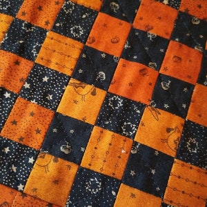 May include: Close-up of a quilted fabric with a checkerboard pattern. Alternating squares are orange and navy blue. The orange squares feature small pumpkin and star patterns. The navy blue squares have star and floral designs. The quilt is likely handmade.