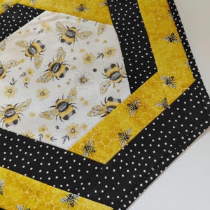 K&ouml;nnte beinhalten: Gesteppter Tischl&auml;ufer mit Bienenmotiv. Die Mitte zeigt einen wei&szlig;en Stoff mit gelben Blumen und gro&szlig;en Hummeln. Der L&auml;ufer ist mit gelbem Wabenmuster und schwarzem Polka-Dot-Stoff eingefasst.