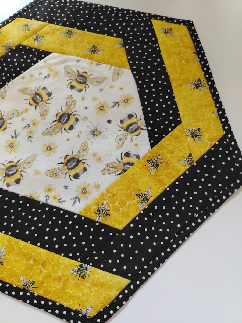 K&ouml;nnte beinhalten: Gesteppter Tischl&auml;ufer mit Bienenmotiv. Die Mitte zeigt einen wei&szlig;en Stoff mit gelben Blumen und gro&szlig;en Hummeln. Der L&auml;ufer ist mit gelbem Wabenmuster und schwarzem Polka-Dot-Stoff eingefasst.