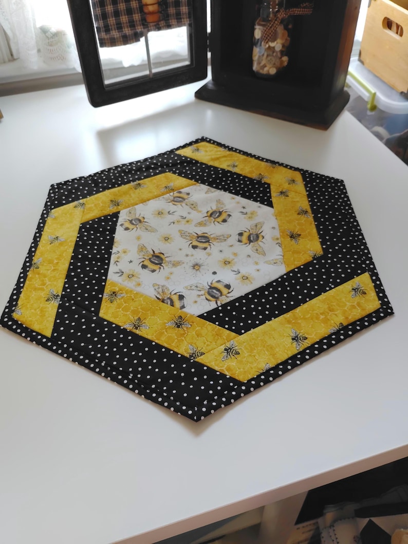 K&ouml;nnte beinhalten: Sechseckiger, gesteppter Tischl&auml;ufer mit Bienenmotiv. Der L&auml;ufer hat einen schwarz-wei&szlig;en Polka-Dot-Rand, gelbe Akzente und ein Mittelteil mit Bienen- und Blumenmustern. Ein dekorativer Artikel f&uuml;r K&uuml;che oder Esszimmer.