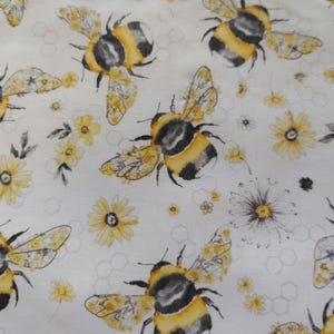 K&ouml;nnte beinhalten: Wei&szlig;er Stoff mit einem sich wiederholenden Muster aus Hummeln, gelben Blumen und Wabenformen. Die Bienen sind schwarz und gelb, und die Blumen sind gelb mit wei&szlig;en Bl&uuml;tenbl&auml;ttern. Der Stoff hat ein skurriles, von der Natur inspiriertes Design.