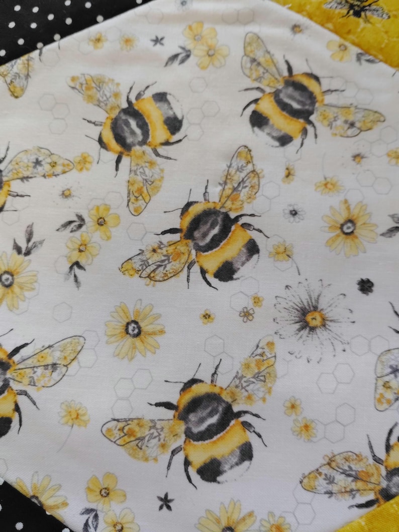 K&ouml;nnte beinhalten: Wei&szlig;er Stoff mit einem sich wiederholenden Muster aus Hummeln, gelben Blumen und Wabenformen. Die Bienen sind schwarz und gelb, und die Blumen sind gelb mit wei&szlig;en Bl&uuml;tenbl&auml;ttern. Der Stoff hat ein skurriles, von der Natur inspiriertes Design.