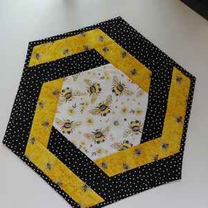 K&ouml;nnte beinhalten: Sechseckiger, gesteppter Tischaufleger mit Bienenmotiv. Das Design zeigt eine wei&szlig;e Mitte mit Bienen und gelben Blumen, umgeben von gelbem Waben- und schwarzen Tupfenstoff. Der Aufleger hat einen Durchmesser von etwa 30 cm.