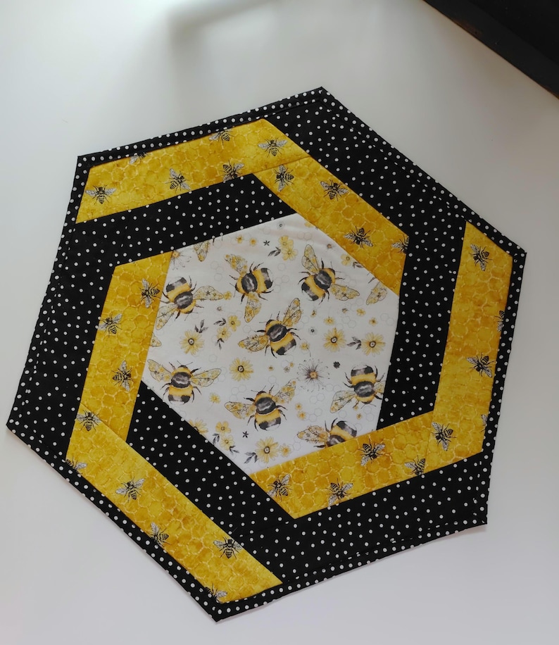 K&ouml;nnte beinhalten: Sechseckiger, gesteppter Tischaufleger mit Bienenmotiv. Das Design zeigt eine wei&szlig;e Mitte mit Bienen und gelben Blumen, umgeben von gelbem Waben- und schwarzen Tupfenstoff. Der Aufleger hat einen Durchmesser von etwa 30 cm.