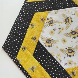 K&ouml;nnte beinhalten: Ein sechseckiger, gesteppter Tischl&auml;ufer mit Bienenmotiv. Das Design umfasst schwarz-wei&szlig;e Punkte, gelbe Wabenmuster und einen wei&szlig;en Stoff mit Bienen- und Blumenillustrationen. Der L&auml;ufer ist ein dekoratives Element f&uuml;r einen Tisch.