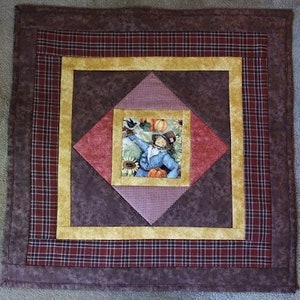 May include: Square quilted wall hanging with a central image of a scarecrow surrounded by pumpkins and sunflowers. The design features a red and gold color scheme with a plaid border and a brown background.