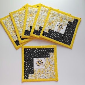 May include: A set of six square fabric coasters. Each coaster features a yellow floral pattern, a black and white polka dot pattern, and a bee illustration. The coasters are trimmed with a bright yellow border.