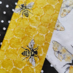 K&ouml;nnte beinhalten: Nahaufnahme eines Stoffes mit einem Bienen- und Wabenmuster. Der Stoff hat ein gelbes Wabenmuster mit schwarzen und wei&szlig;en Bienen. Angrenzend ein wei&szlig;er Stoff mit einer Biene und ein schwarzer Stoff mit wei&szlig;en Punkten.