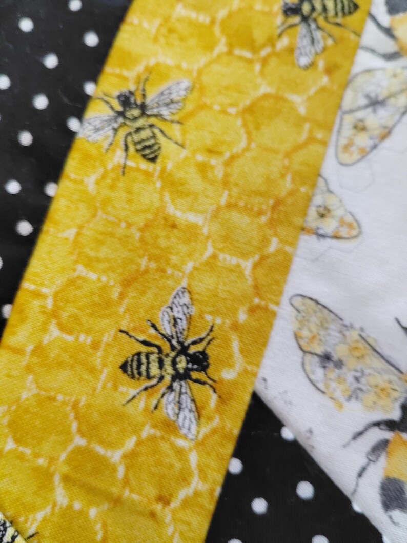 K&ouml;nnte beinhalten: Nahaufnahme eines Stoffes mit einem Bienen- und Wabenmuster. Der Stoff hat ein gelbes Wabenmuster mit schwarzen und wei&szlig;en Bienen. Angrenzend ein wei&szlig;er Stoff mit einer Biene und ein schwarzer Stoff mit wei&szlig;en Punkten.